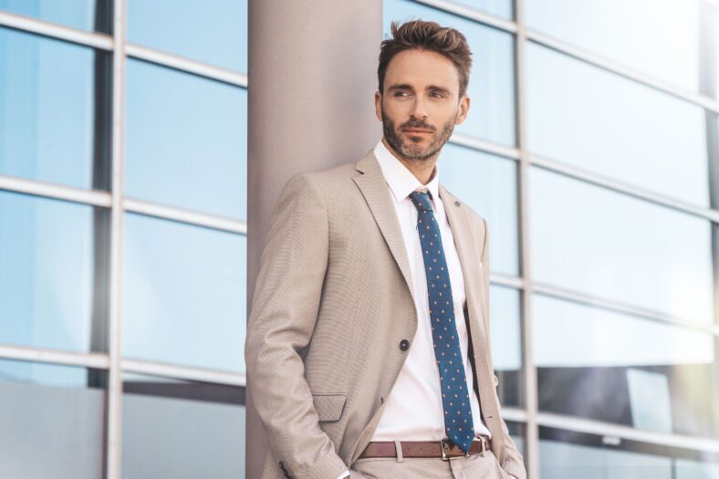 Young, handsome, elegant businessman is standing by the glassy office building wearing a light suit