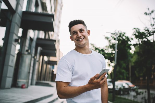 Happy hipster guy in casual t-shirt with copy space for brand label standing at urban setting and laughing during online communication via cellular gadget, positive cheerful man smiling outdoors