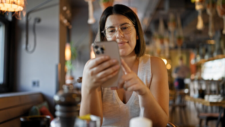 Young beautiful hispanic woman using smartphone sitting on the table at the restaurant