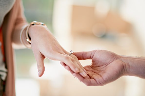 Couple, hands and engagement with ring in home, love and relationship with diamond jewellery on fiancee. Affection, wedding proposal and commitment with partner, romance and apartment together.