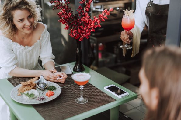 Charming blond lady looking at young man with smile while bartender holding glass of cocktail. Couple sitting at the table with food, bouquet of reed berries and smartphones