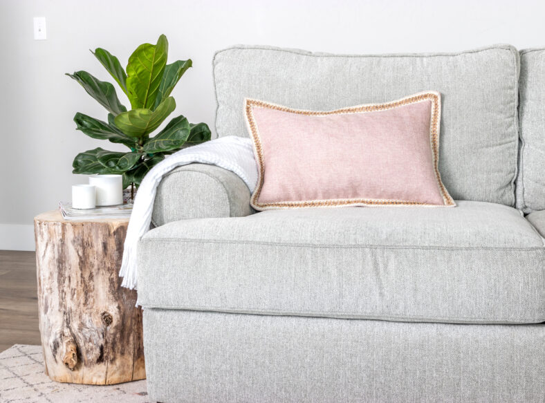 A modern farmhouse home decor scene with couch, wood stump end table, throw pillows and fiddle leaf fig houseplant.