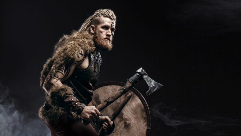 Medieval warrior berserk Viking with tattoo on skin, red beard and braids in hair with axe and shield attacks enemy. Concept historical photo