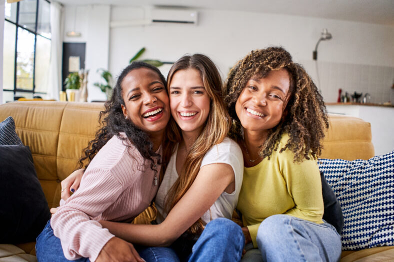 Portrait of three attractive female room mates looking smiling at camera. Group of cute cheerful girls hugging sitting on couch at home. Happy friends posing for photo indoors. Generation z and rent.