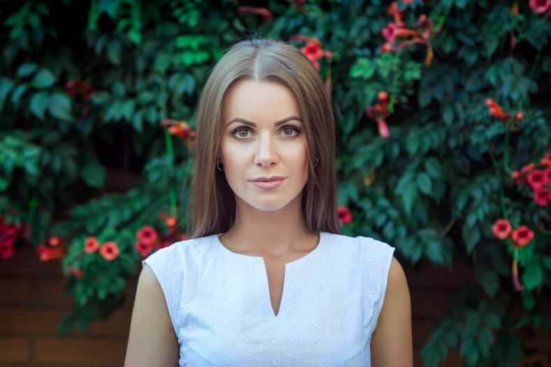 Portrait of beautiful young serious woman, wearing elegant white dress, standing in front of brick wall covered by blooming trumpet creeper plant.
