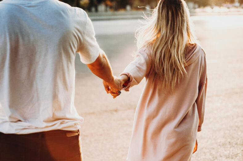 Crop back view of casual couple holding hands and walking on street in sunshine