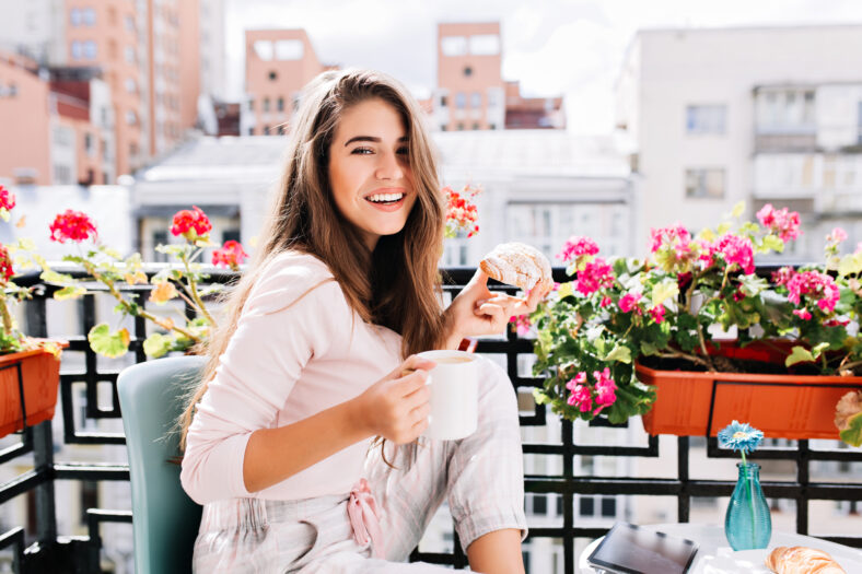Portrait pretty girl having breakfast on balcony surround flowers in the sunny morning in city. She holds a cup, croissant, smiling to camera