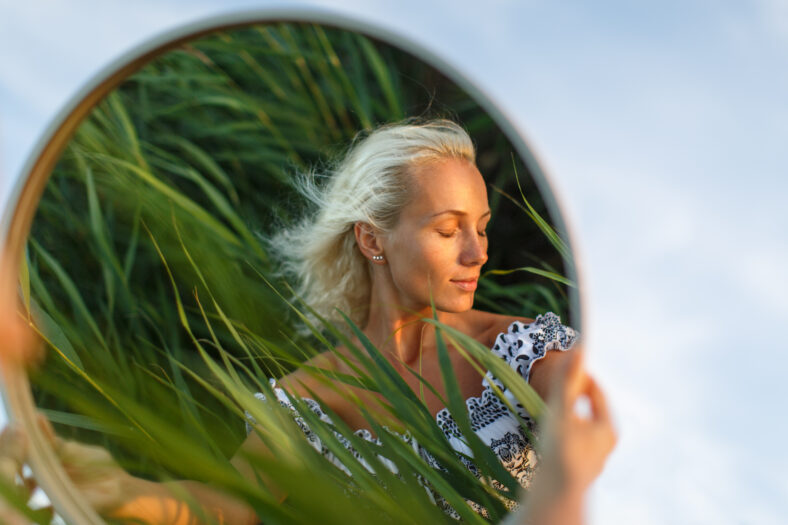 Pretty blonde woman with curly hair in dress holding a round mirror at sunset, closed eyes, green reed on background. Reflection of female in the mirror