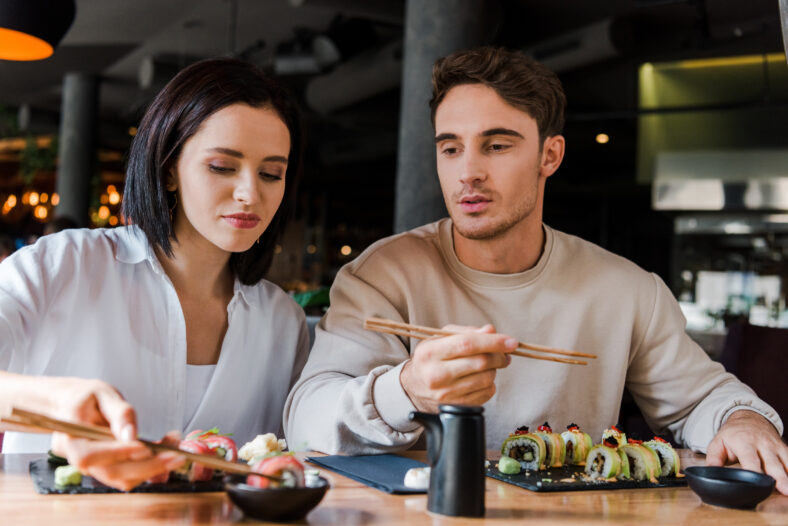 selective focus of attractive woman holding chopsticks with tasty sushi near man in restaurant