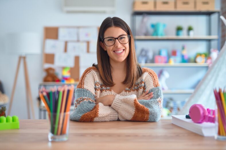 Young beautiful teacher woman wearing sweater and glasses sitting on desk at kindergarten happy face smiling with crossed arms looking at the camera. Positive person.