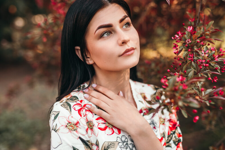 High angle of stylish young lady looking up and thinking while standing near tree with red flowers on sunny day in spring garden