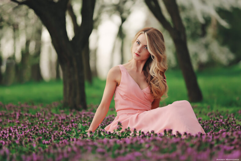 Beautiful blonde young woman in colorful flowers. girl with make-up and hairstyle in pink dress in blossoming spring park. Art work of romantic woman .Pretty tenderness model looking at the camera.