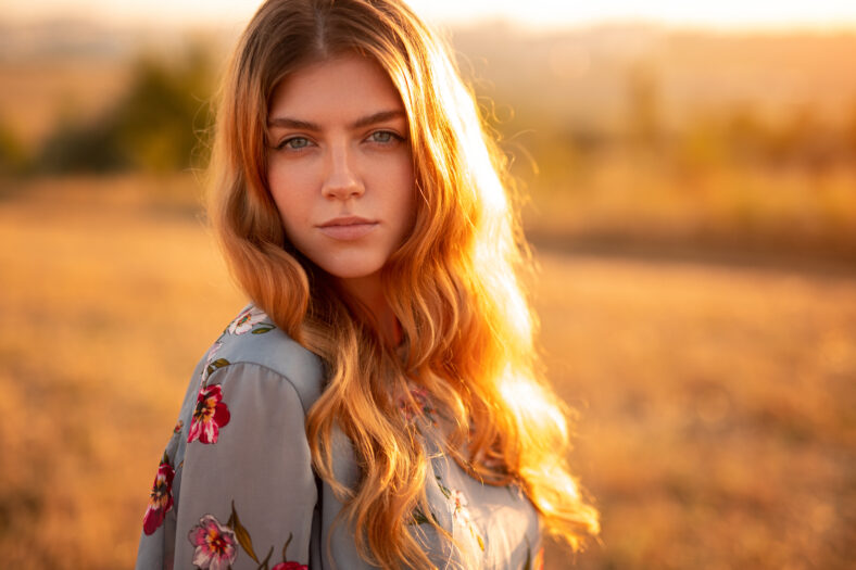 Charming young redhead female in stylish dress looking at camera while standing in yellow field in sunny autumn day