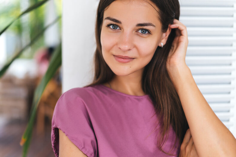 Close-up portrait of beautiful young brunette woman with amazing green eyes in modern white style cafe. Girl in pink dress posing near bright wooden wall, she has natural make-up