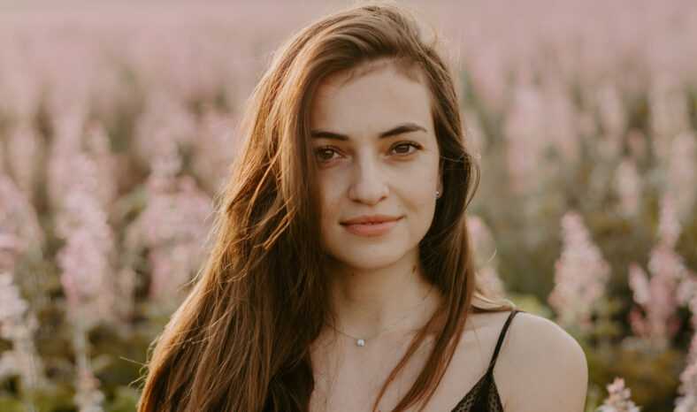 Portrait of a young woman in a flower field looking at camera with neutral expression, backlist by sunset golden light