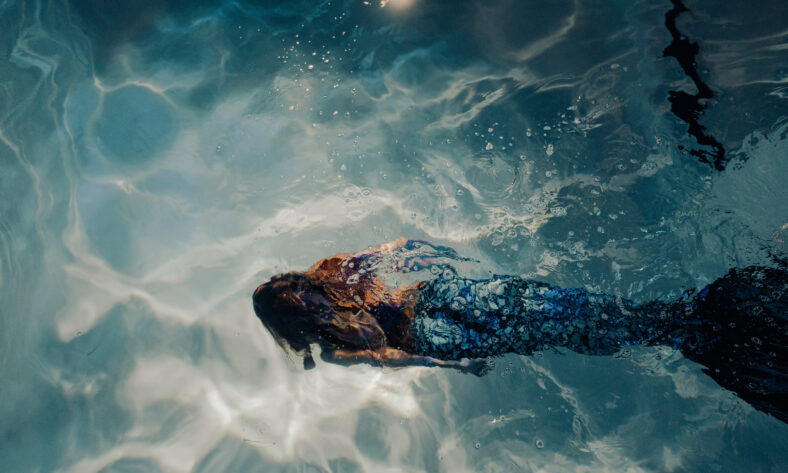 Beautiful young woman with mermaid tail swims and dives under sunlit deep water.