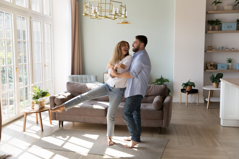 Spouses listen music dance barefoot in fashionable warm sunny living room, full length. Happy homeowners couple in love enjoy romantic date feel enamoured. New home, bank loan for young family concept