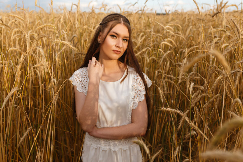Portrait of tranquil woman with long fair hair in white dress standing in wheat field in summer in rural area