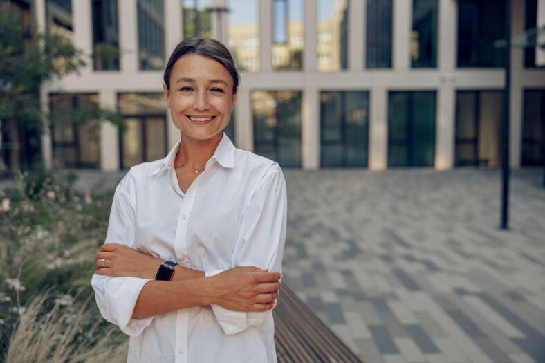 Portrait of smiling female manager standing on modern building background and looks camera