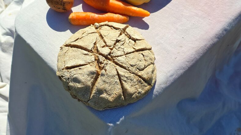 A rustic, round loaf of bread with a star-like scoring pattern sits on a white cloth alongside carrots and a potato, evoking a simple, historical meal setting.