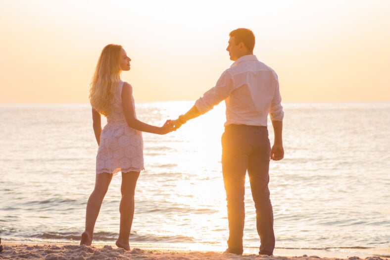 Beautiful young romantic couple holding hands on seaside in rays of rising sun. Two happy lovers at golden sunrise. Warm colors.