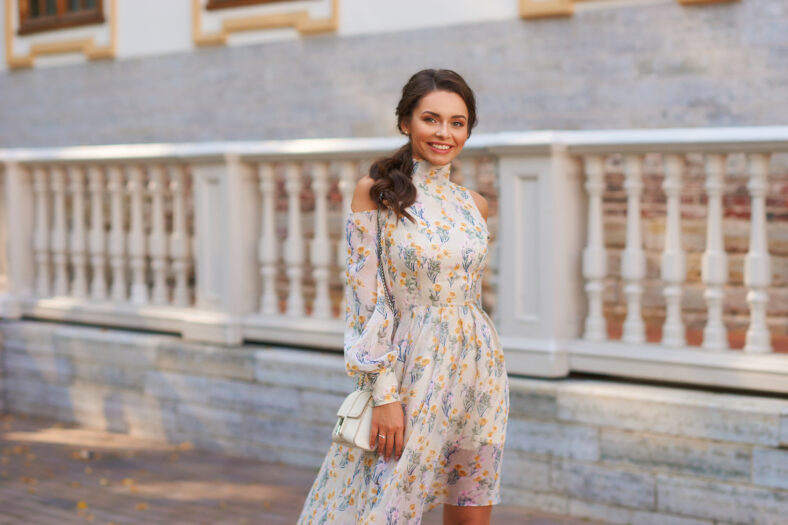 Pretty smiling young woman with long brunette hair wearing fashionable floral dress standing on street against balustrade on background. Happy female model in elegant stylish outfit posing outdoors.