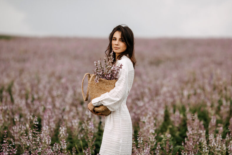 Young woman in white summer dress, standing in a big field with sage in bloom, holding a wicker bag with flowers.