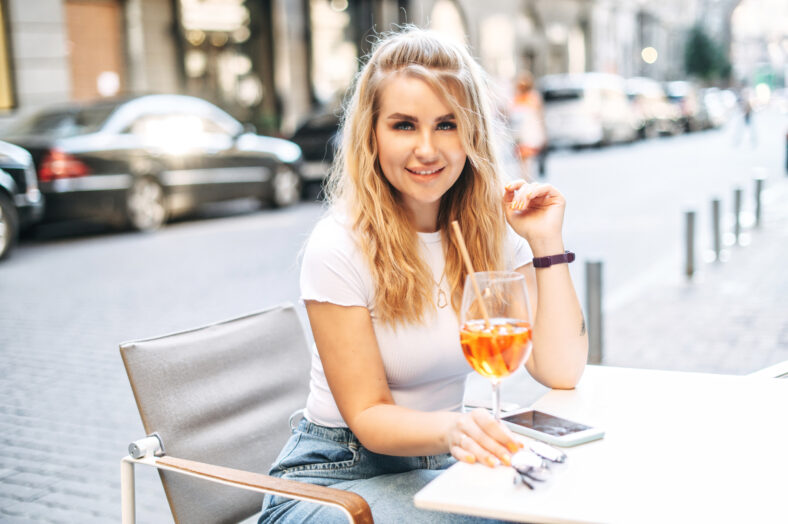 Beautiful blonde girl with a cocktail in hands in the outdoor cafe, woman holds elegant glass with drink and looks at camera with a smile