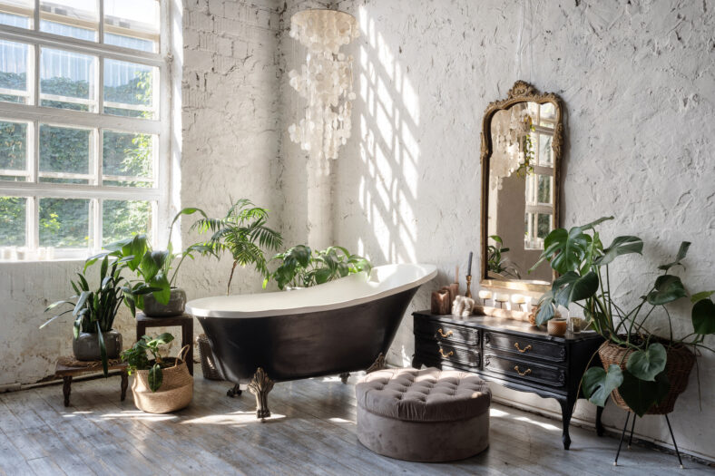 Side view of white cozy bathroom with black tub, wooden classic commode, mirror, decor, plants and interior design in boho chic style. Concept of comfortable and decorated room at home