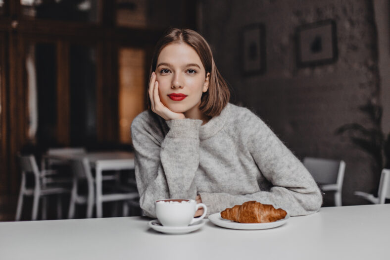 Brown-eyed girl in woolen sweater leaned on white table in cafe and looking at camera. Photo of woman with red lips ordering coffee and croissant