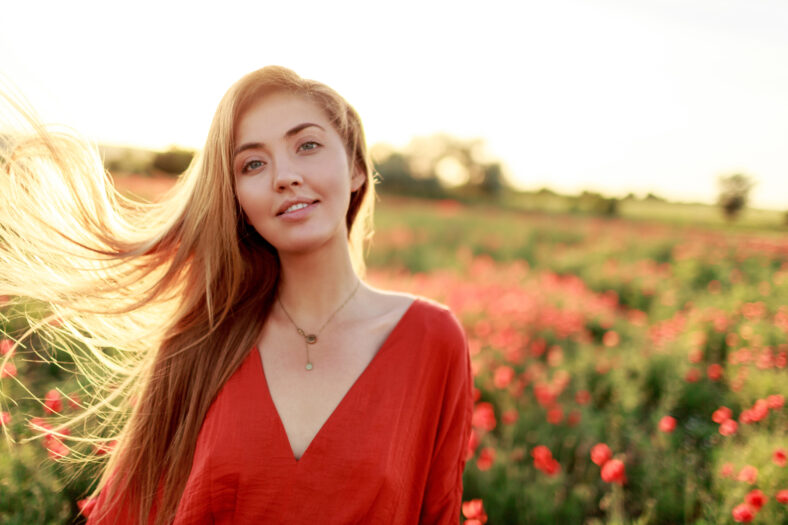Graceful blonde woman looking in camera. Windy hairs. Picturesque poppy field in warm sunset colors.
