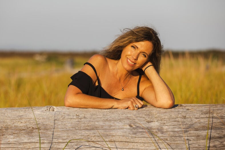 Pretty middle aged woman smiles while leaning on driftwood log outside in nature during the golden hour