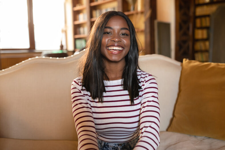 Positive emotions. Portrait of cheerful African American woman smiling, laughing and looking at camera, sitting on couch at home. Young black female expressing joy and happiness indoors