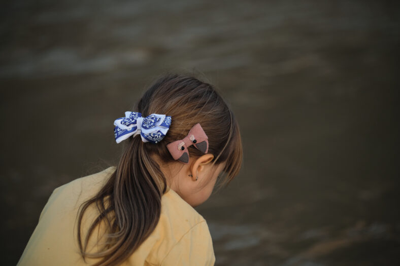 Hairstyle of Caucasian little girl close up - brown hair gathered in tail and two bows blue and pink. Beautiful hairstyle of child is little side view from behind.