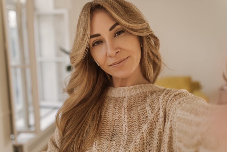 Pretty caucasian adult woman makes selfie imposingly looking at camera in light room. Blonde girl with long hair wears warm sweater. Lifestyle, female beauty concept