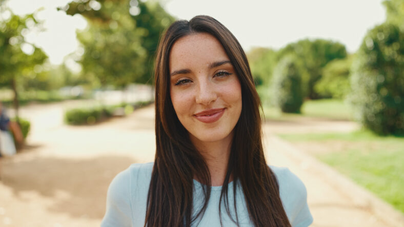 Close-up of young smiling woman with long brown hair wearing white crop top posing for the camera in the park . The girl opens her eyes and smiles at the camera