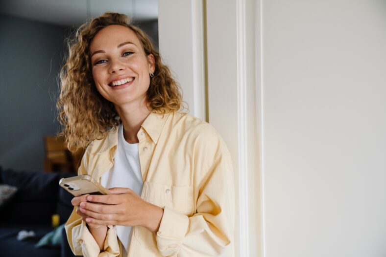 Young beautiful attractive smiling happy curly woman holding phone and looking at camera while standing at home