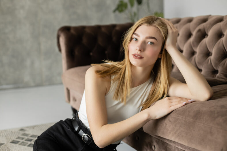 Portrait of young blonde woman looking at camera near couch at home,stock image