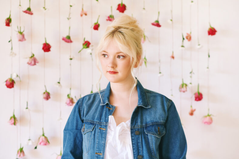 Studio portrait of pretty young teenage 15 - 16 year old girl wearing denim jacket, posing on white background with hanging flowers, beauty and fashion concept