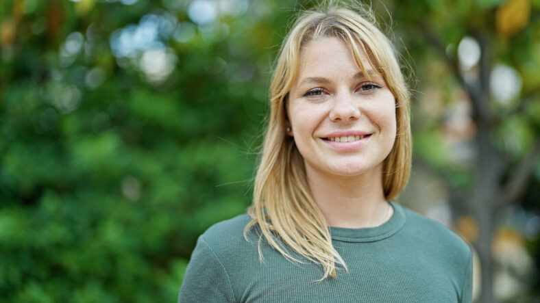 Joven rubia sonriendo con confianza de pie en el parque