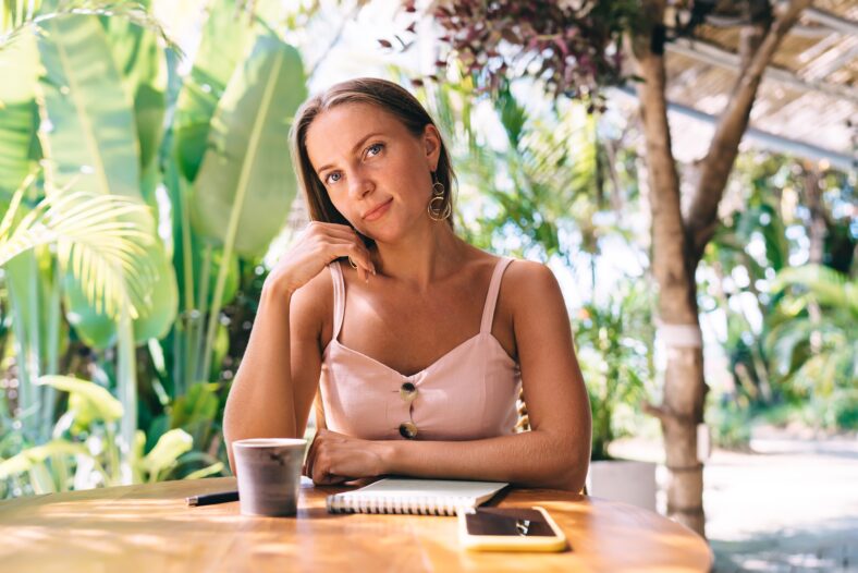 Content female in casual summer outfit looking at camera while sitting with cup of coffee and smartphone at table in cafe