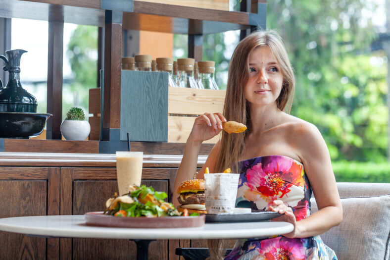 Casual lunch setting with delighted woman, vibrant dress, indulging in burger and salad, serene restaurant environment. Everyday healthy eating.
