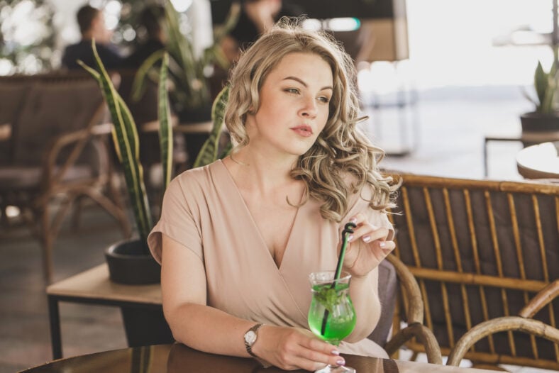 A young blonde woman plus size model sits with a cocktail on the summer area of a city cafe.