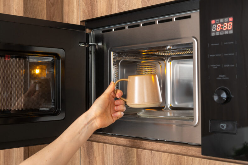 A person's hand reaches into a microwave to place inside a ceramic mug with cold beverage to heat coffee at home or work