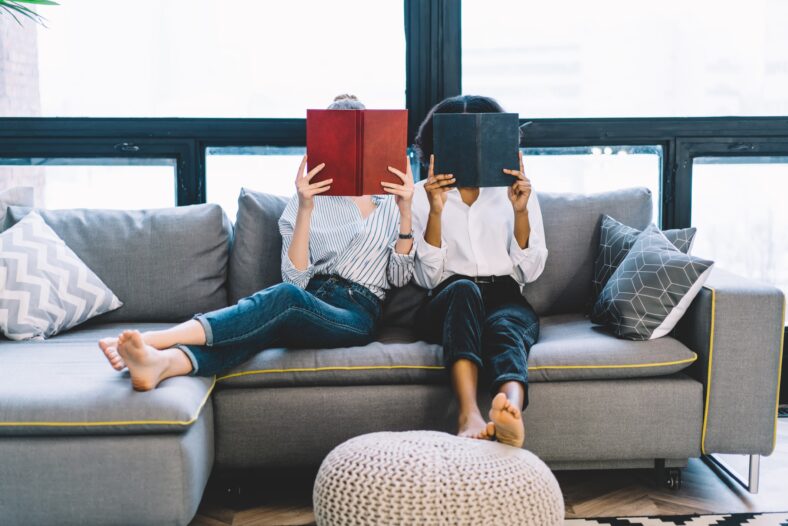 Two multicultural students sitting on comfortable couch and holding literature books in front of faces spending time together at home on weekend, diversity young women reading interesting bestsellers