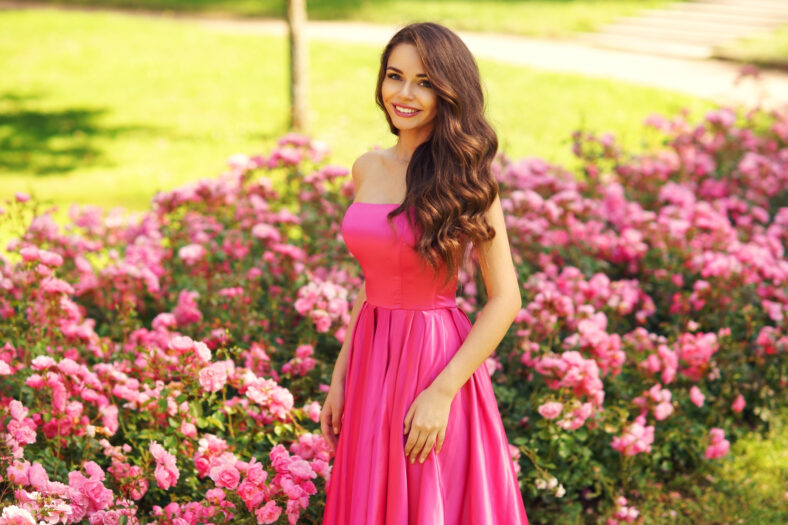 Princess. Young beautiful pretty woman posing in long evening luxury dress against bushes with pink roses on a sunny summer day.