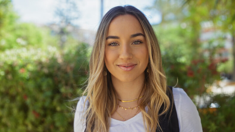 Attractive young adult blonde woman in an urban park with greenery, smiling outdoors on a sunny day.