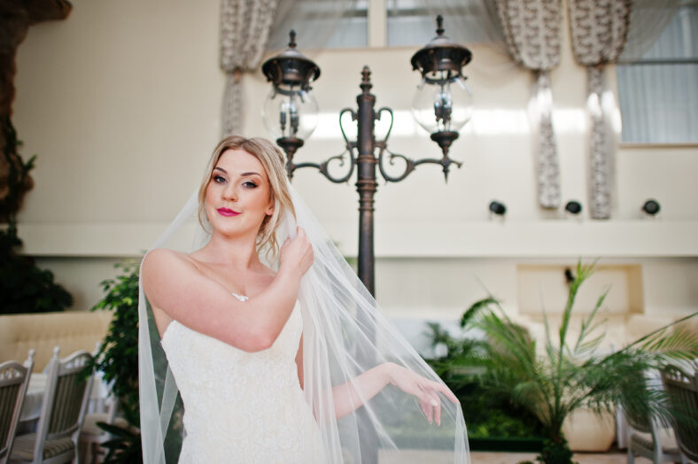 Gorgeous blonde bride with long veil posed at great awesome wedding hall background lantern