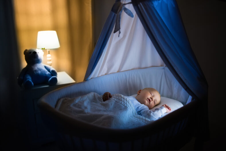 Adorable baby sleeping in blue bassinet with canopy at night. Little boy in pajamas taking a nap in dark room with crib, lamp and toy bear. Bed time for kids. Bedroom and nursery interior.