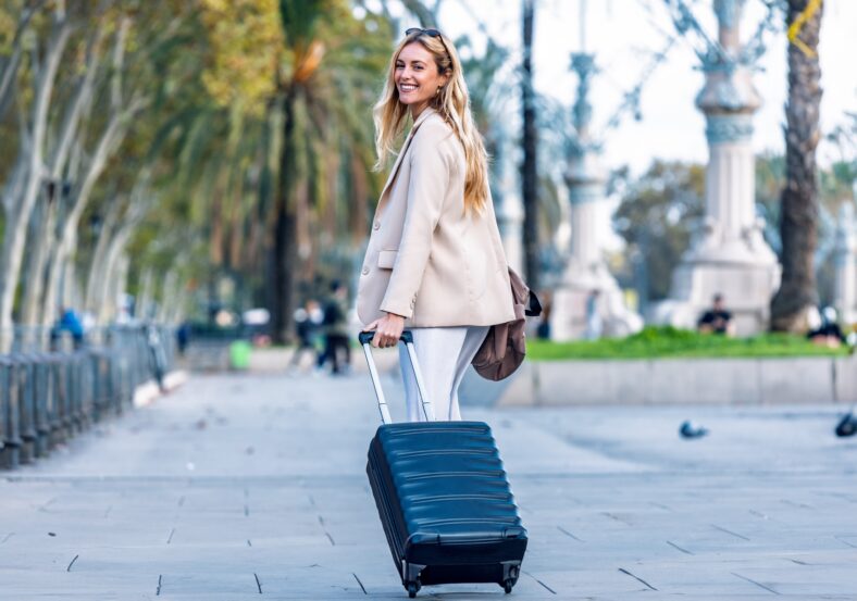 Shot of pretty young woman walking with suitcase while looking at camera through the city